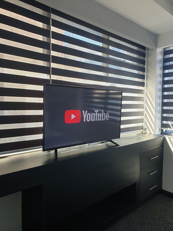 Une chambre moderne dotée d’un téléviseur à écran plat fixé à un bureau sombre, d’une fenêtre avec des stores et d’un sol recouvert de moquette.