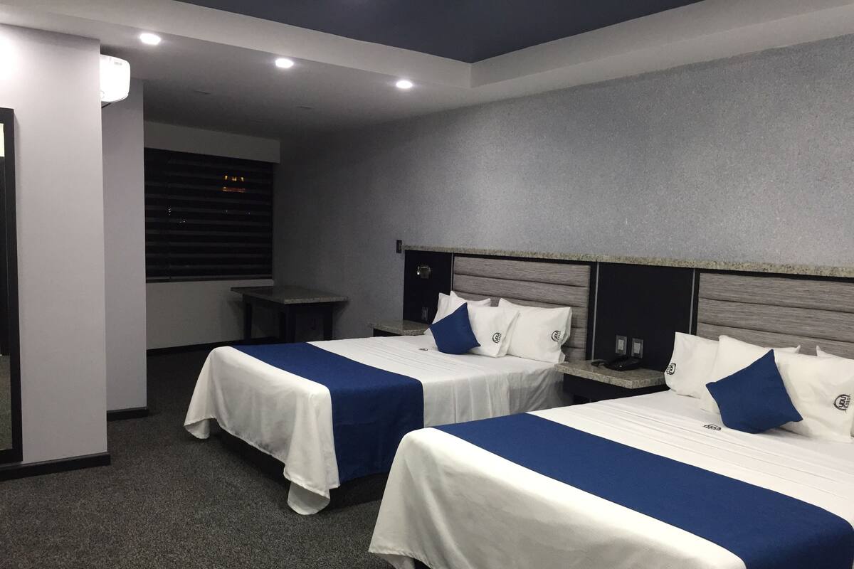 A hotel room with two beds, each with a white and blue bedspread, a headboard, and a bedside table with a lamp.