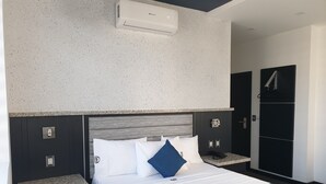 Une chambre d’hôtel comprenant un lit, une table de chevet, un climatiseur fixé au mur, un espace douche et une porte avec une poignée.