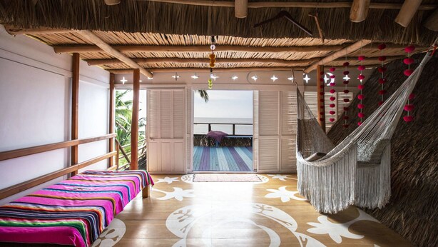 Una habitación con una hamaca, una cama con una manta de colores, un suelo de madera con motivos florales y un techo de paja.