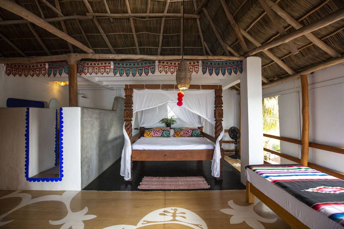 Una habitación con una cama de madera, una mesita de noche pequeña, una silla, una ventana y un techo de paja.