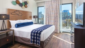 Una habitación de hotel moderna con una cama grande, ventilador de techo, vista a la ciudad y un escritorio con televisor de pantalla plana.