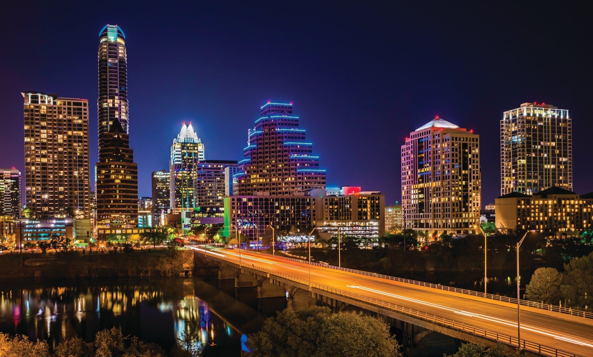 A city skyline at night with illuminated buildings, a river, and a bridge.