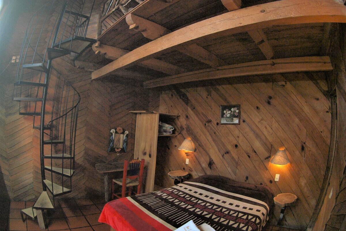 Une pièce rustique avec un escalier en colimaçon, un lit avec une literie à motifs, deux lampes fixées au mur et une petite table avec une chaise.