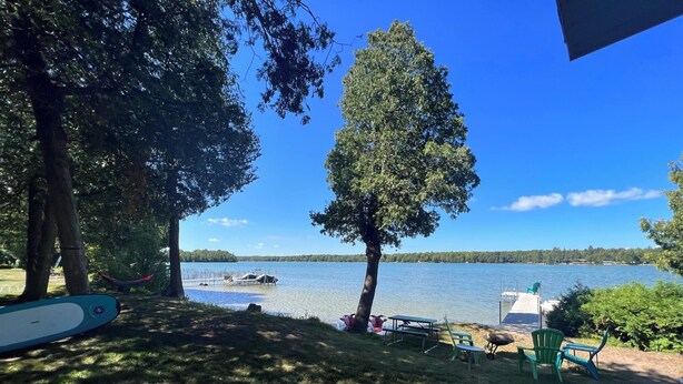 A lakeside view with picnic tables and chairs, a hammock, and a dock with boats. - Fabulous 2 Bed, 2 Bath Cottage On Kangaroo Lake (Baileys Harbor)