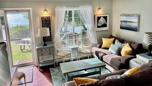 A cozy living room with a ceiling fan, a sofa with cushions, a coffee table, and a view of the outdoors through a glass door. - Fabulous 2 Bed, 2 Bath Cottage On Kangaroo Lake (Baileys Harbor)