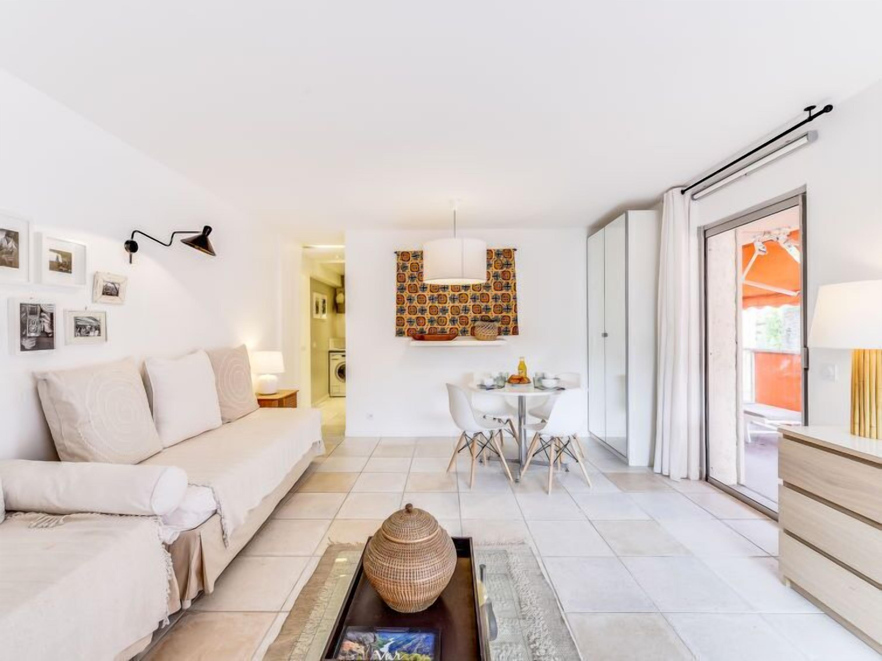A modern living room with a sofa, coffee table, and dining area. There is a wall-mounted light fixture and a patterned wall hanging.