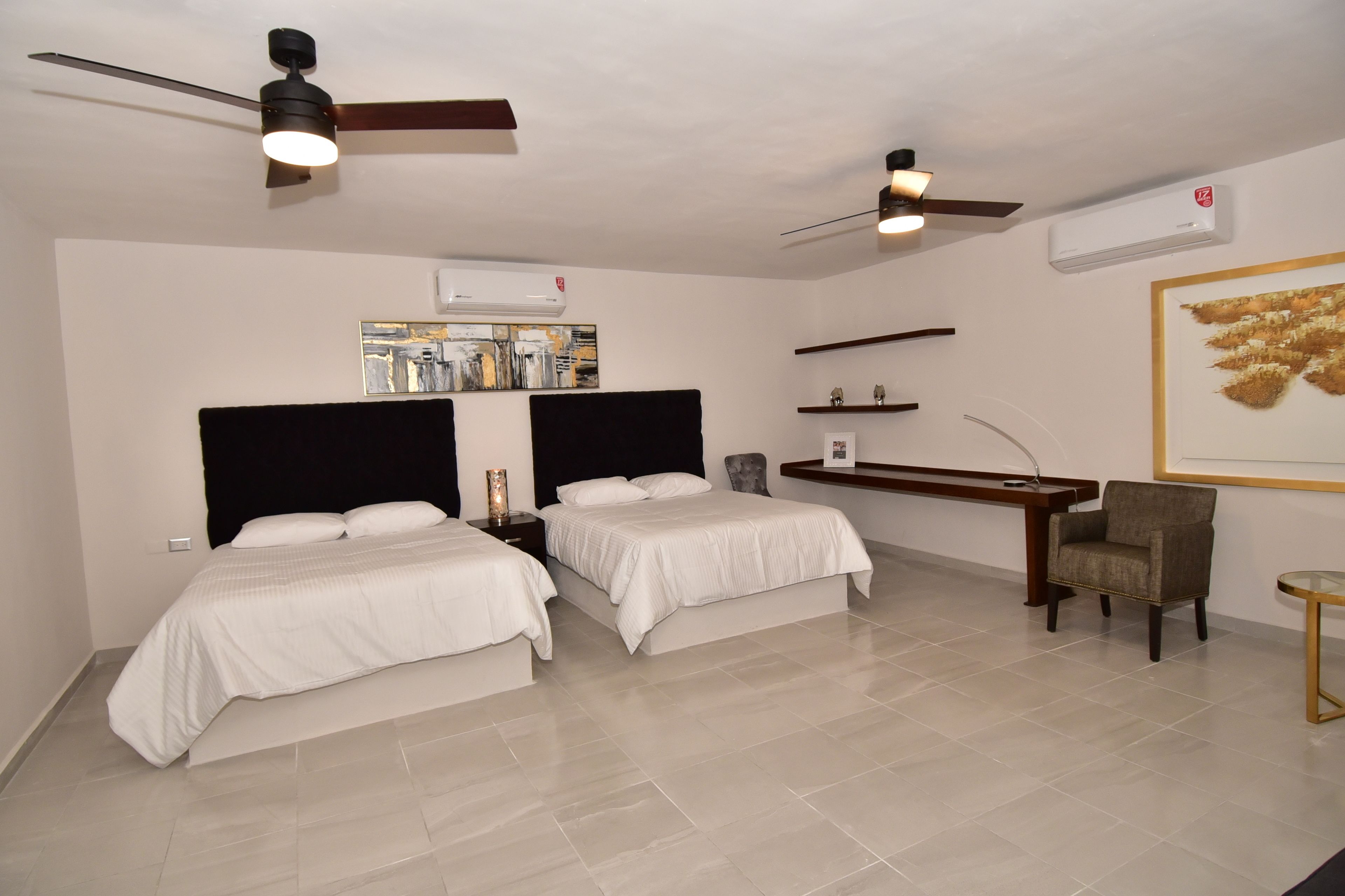 Une chambre d’hôtel avec deux lits, un bureau, une chaise, des ventilateurs de plafond et des œuvres d’art accrochées aux murs.
