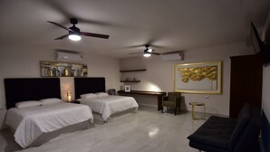 Une chambre d’hôtel avec deux lits, un bureau, une chaise, un ventilateur de plafond et des œuvres d’art accrochées aux murs.