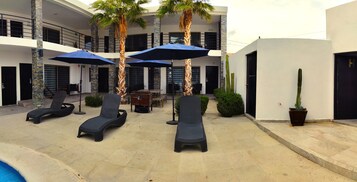 Zona junto a la piscina con sillones de descanso, sombrillas y palmeras.