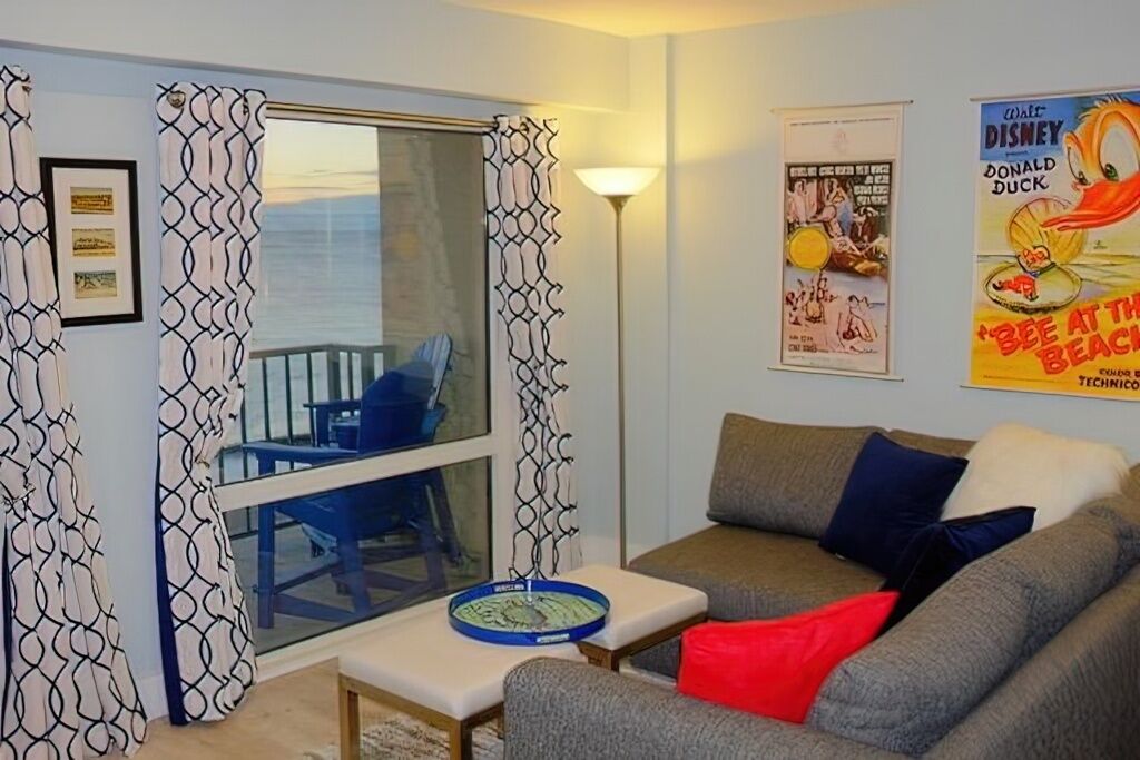 Un salon avec un canapé, une table basse et une fenêtre avec des rideaux. Il y a des affiches au mur et une lampe sur une table d’appoint.
