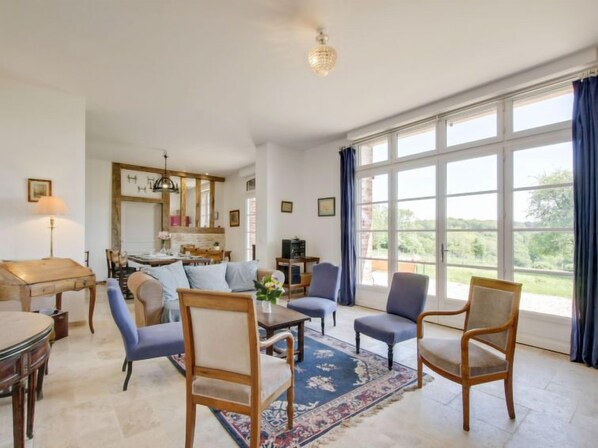 A spacious living room with a large window, a dining area, and a kitchen in the background.
