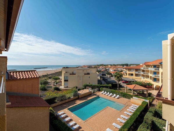 A pool area with lounge chairs, a clear view of the beach, and a building with balconies. - Piscine Extérieure ! Abordable + Cuisine Équipée (Valras-Plage)