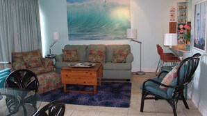 A living room with a sofa, coffee table, and chairs, a painting of a wave on the wall, and a bookshelf with books and decorative items. - Ocean View from the Balcony! (Rehoboth Beach)