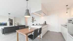 A modern kitchen with a central island, a dining area with a wooden table and chairs, and a living area with a black sofa and a TV.