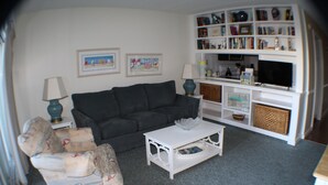 A living room with a dark grey sofa, a white coffee table, patterned armchairs, a bookshelf, and a flat-screen TV.