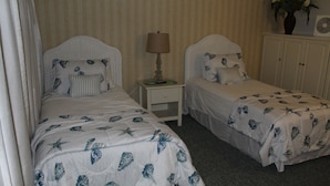 Two single beds with matching headboards and footboards, a bedside table with a lamp, a nightstand, and a cabinet with a potted plant.