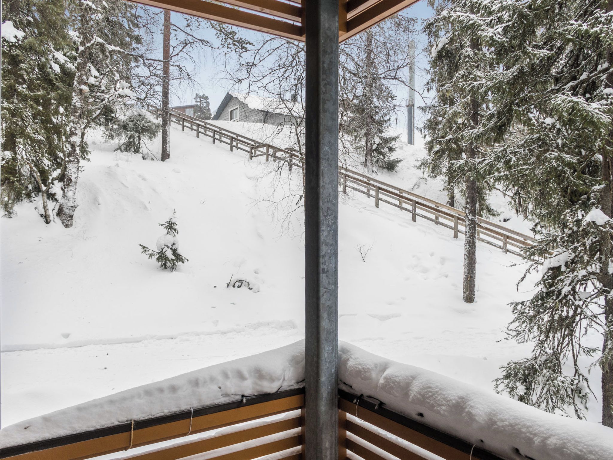 Área externa nevada com deck de madeira, árvores cobertas de neve e corrimão de metal.