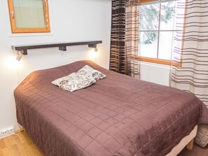 A bed with a brown quilt, a bedside lamp, and a window with striped curtains. - Karhunvartijan linna a3 by Interhome (Kuusamo)