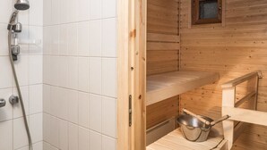 Interior de una cabaña de madera con zona de ducha, estantes de madera y un cubo de metal.