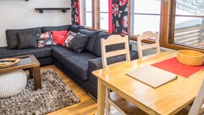 Una sala de estar acogedora con un sofá gris oscuro, una mesa de comedor de madera y un tapete rojo. Hay dos ventanas con cortinas estampadas y una estantería contra la pared.
