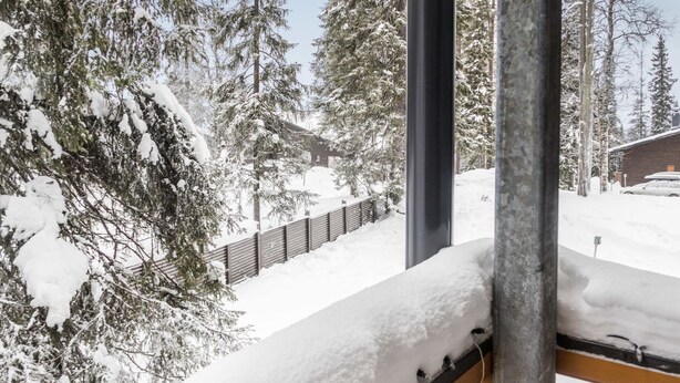 Escena nevada al aire libre con un banco de madera, árboles cubiertos de nieve y un edificio de ladrillo al fondo.