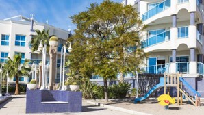 A beachfront hotel with a children's play area, palm trees, and a decorative fountain.
