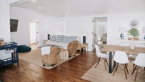 A spacious living area with a wooden floor, a white sofa with patterned cushions, wicker chairs, a dining table with white chairs, and a small side table. - Historic 3 bedroom Cottage for 8 with pool -walk to the beach! (Tybee Island)