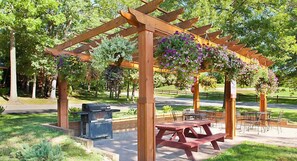 Una pergola in legno con cesti di fiori appesi, tavoli da picnic e una griglia barbecue in un parco.