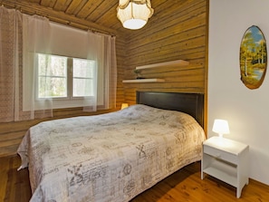 A bedroom with a wooden ceiling, a bed with a patterned bedspread, a nightstand with a lamp, and a window with curtains. - Honkaniemi by Interhome (Joensuu)