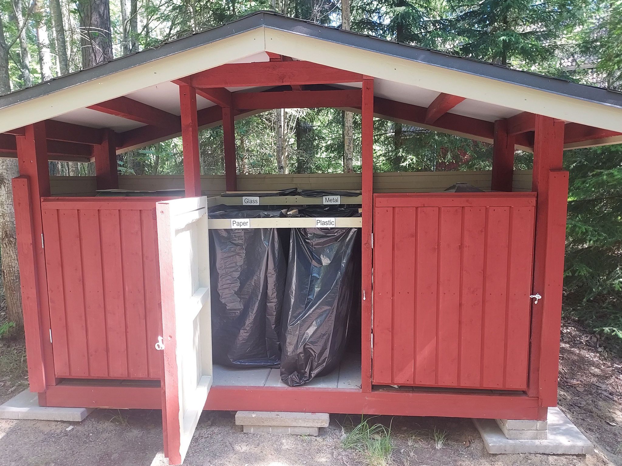 A red outdoor structure with a roof, featuring a trash bin with labeled compartments for glass, metal, paper, and plastic.