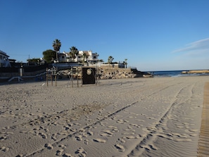 A beach with footprints, a wooden boardwalk, and a building with palm trees in the background.