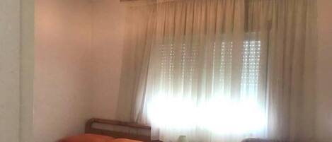 A room with two single beds, each with a striped bedspread and an orange pillow. There is a small bedside table with a lamp on one side and a drawer on the other. The room has a window with sheer curtains and a light fixture above the beds.