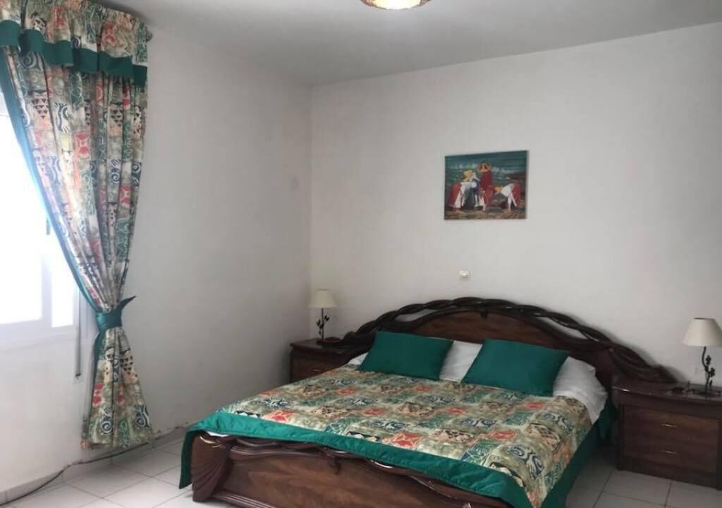 A bedroom with a wooden bed, green bedding, a patterned curtain, and a small painting on the wall.