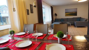 A dining area with a table set for four, red placemats, and wine glasses. There is a sofa and a painting in the background. - Sun-drenched first floor apartment on the dyke (Puttgarden)