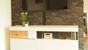 A modern living room with a flat-screen TV mounted on a textured stone wall, a white entertainment unit with built-in storage, and a small potted plant on the unit. - Sun-drenched first floor apartment on the dyke (Puttgarden)