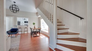A spacious living area with hardwood flooring, a dining table with chairs, a blue armchair, and a staircase with white walls and wooden steps.