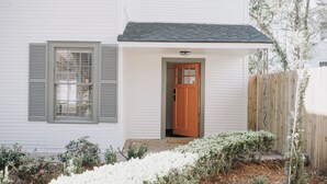 A white house with a wooden door and shutters, a small porch, and a garden with white flowers.