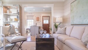 A spacious living room with a sofa, armchair, and coffee table. There is a bookshelf and a door leading to another room. - Broad Creek Cottage in River Dunes (Oriental)