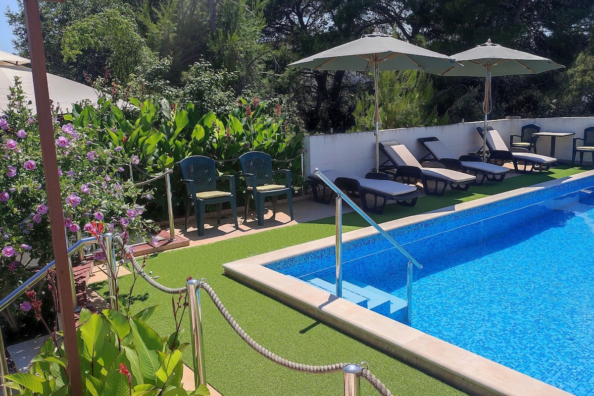 Piscina com borda revestida, cercada por grama sintética verde, e equipada com espreguiçadeiras e guarda-sóis.