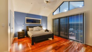 Ein Schlafzimmer mit Holzboden, einem Bett, einem Nachttisch, einem Beistelltisch, einem Fenster und einer Glasschiebetür, die auf einen Balkon führt.