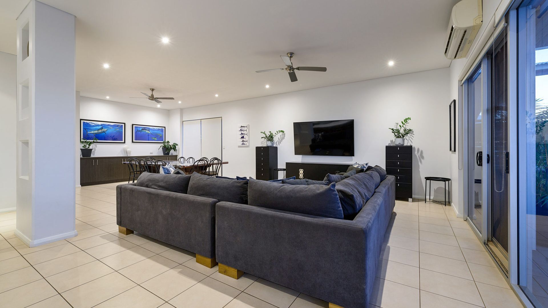 A modern living room with a grey sofa, a dining area with chairs, a TV mounted on the wall, and ceiling fans.
