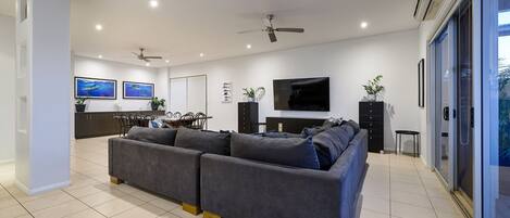 A modern living room with a grey sofa, a dining area with chairs, a TV mounted on the wall, and ceiling fans.
