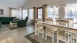Un salon spacieux avec un canapé vert, une table à manger entourée de chaises blanches et de grandes fenêtres donnant sur des arbres couverts de neige.