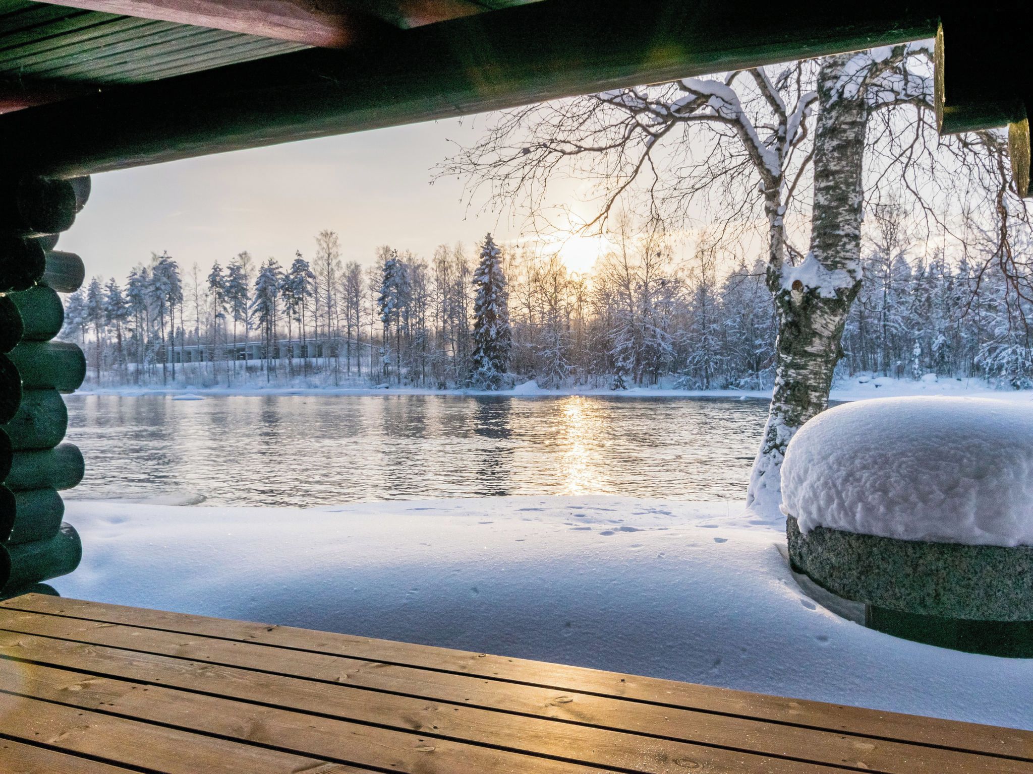 A wooden deck overlooking a snow-covered lake with trees and a cabin in the background.
