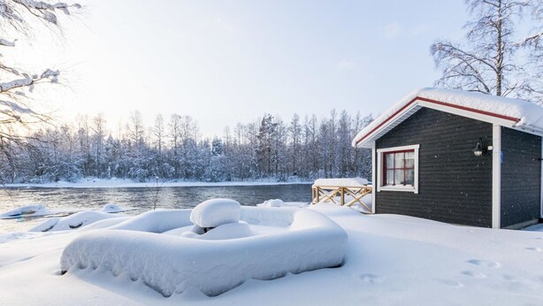 Une cabane dotée d’une petite terrasse et d’un banc, entourée d’arbres couverts de neige et d’une rivière.