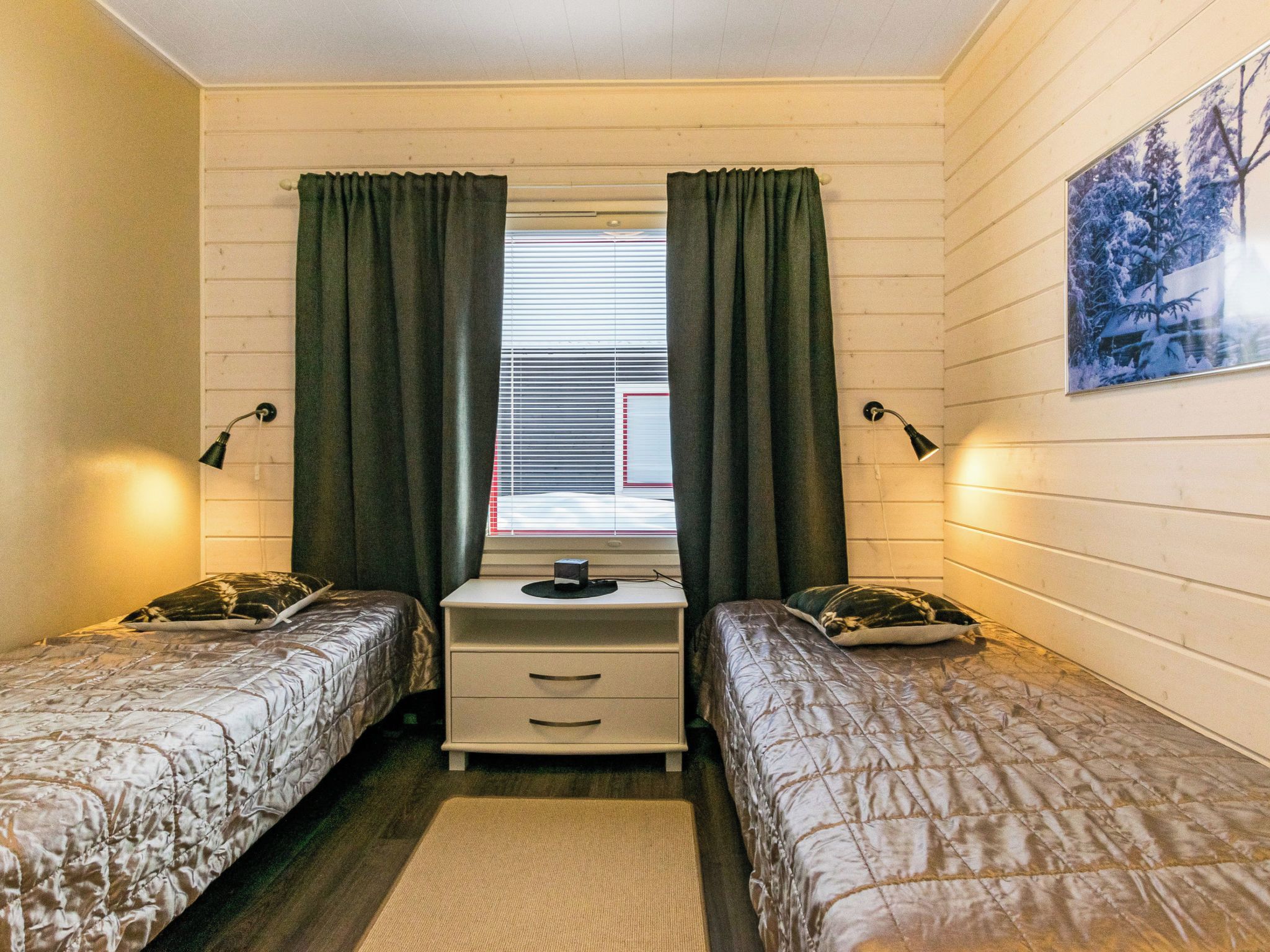A room with two beds, a desk, a window with curtains, wall-mounted lamps, and a framed picture on the wall.