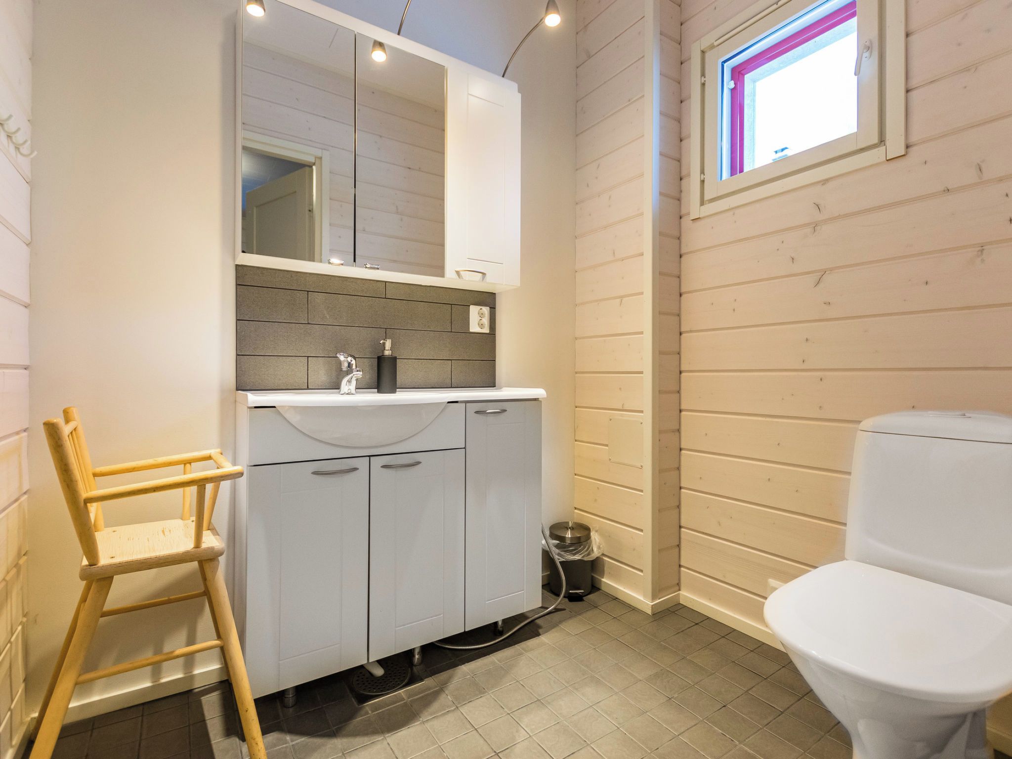 Une salle de bain avec un lavabo, un miroir et des toilettes, tous de couleur blanche. On y trouve également une chaise en bois et une poubelle.