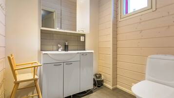 Une salle de bain avec un lavabo, un miroir et des toilettes, tous de couleur blanche. On y trouve également une chaise en bois et une poubelle.