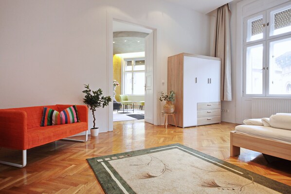 Una sala de estar moderna con piso de madera, un sofá con cojines de colores, una cama, un armario y una ventana con cortinas.